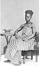  
Code: 0002 4047
Category: People from Elmina

Three portraits of Elmina women, unidentified
2. Lady sitting in chair, formal portrait.

This set of three portraits is not identified by Muller. Possibly he regarded the portraits of women in traditional dress as ethnographic curiosities. The compositions of the portraits indicates, however, that these are photo's made for the use of the depicted persons themselves.
The young woman shows her virginity by showing her bared breasts. A portrait like this was often made in well to-do circles to commemorate the engagement of the girl in question. The group portrait of two women and two girls seems to be a family group of three generations (grandmother, mother, daughters).
